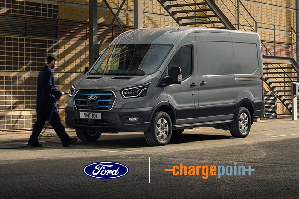 Grey electric van parked in front of a brick warehouse, a man is preparing to charge the vehicle.