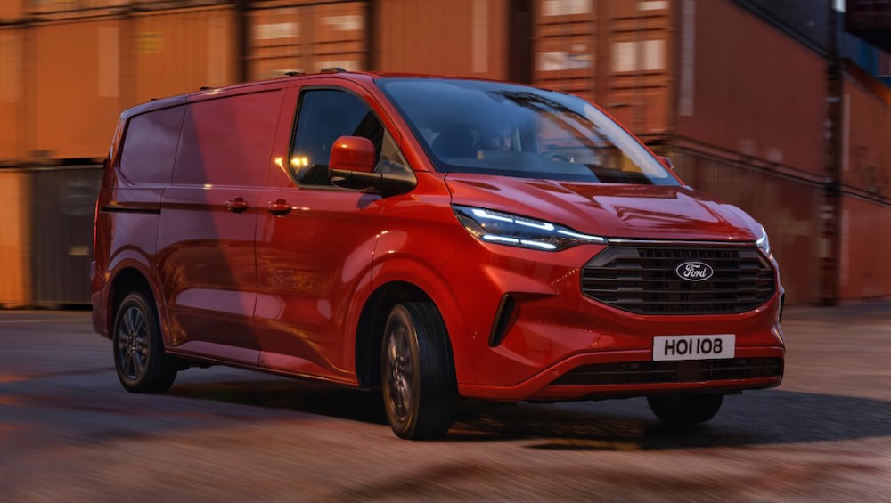 Red Ford Transit Custom van driving at dusk, with blurred shipping containers in the background.