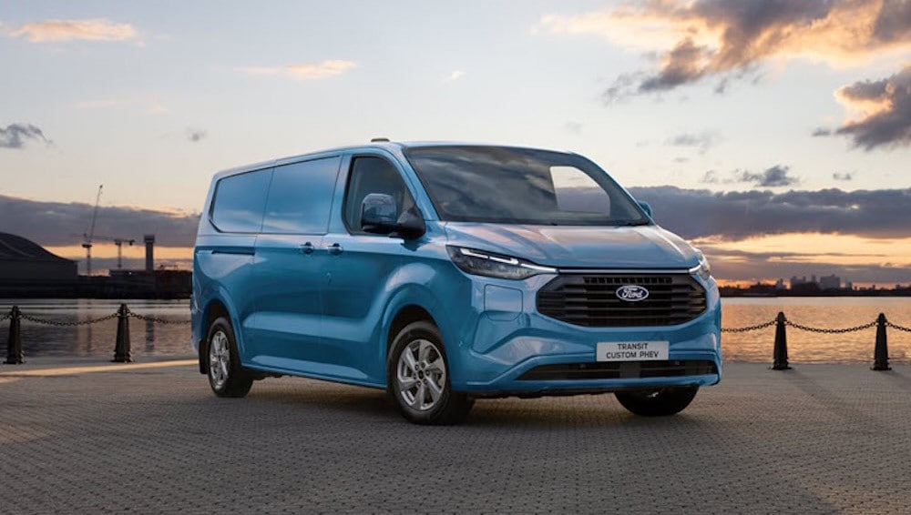 Blue Ford Transit Custom PHEV van parked on a paved area next to a body of water at sunset.