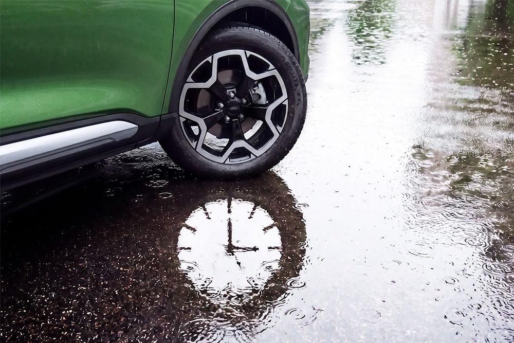 A green Ford Kuga's front wheel sits next to a puddle, with a clock face reflected in the water's surface.