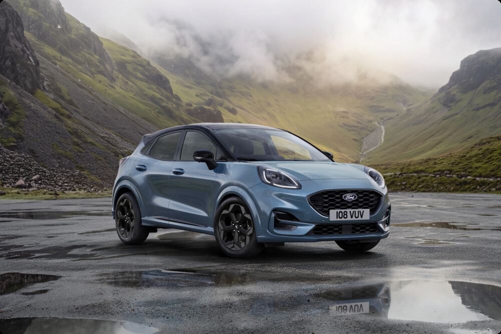 A light blue Ford Puma parked on a wet road, seen from a front three-quarter angle in a mountain landscape.