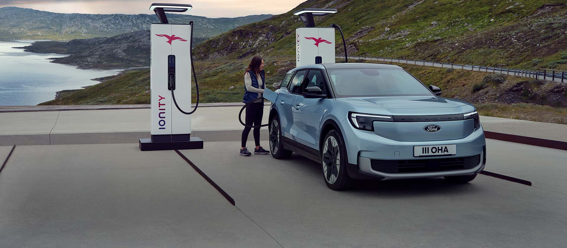 A woman plugs a blue, all-electric Ford Explorer into a public charging station using a cable. The car is parked by mountains and water.