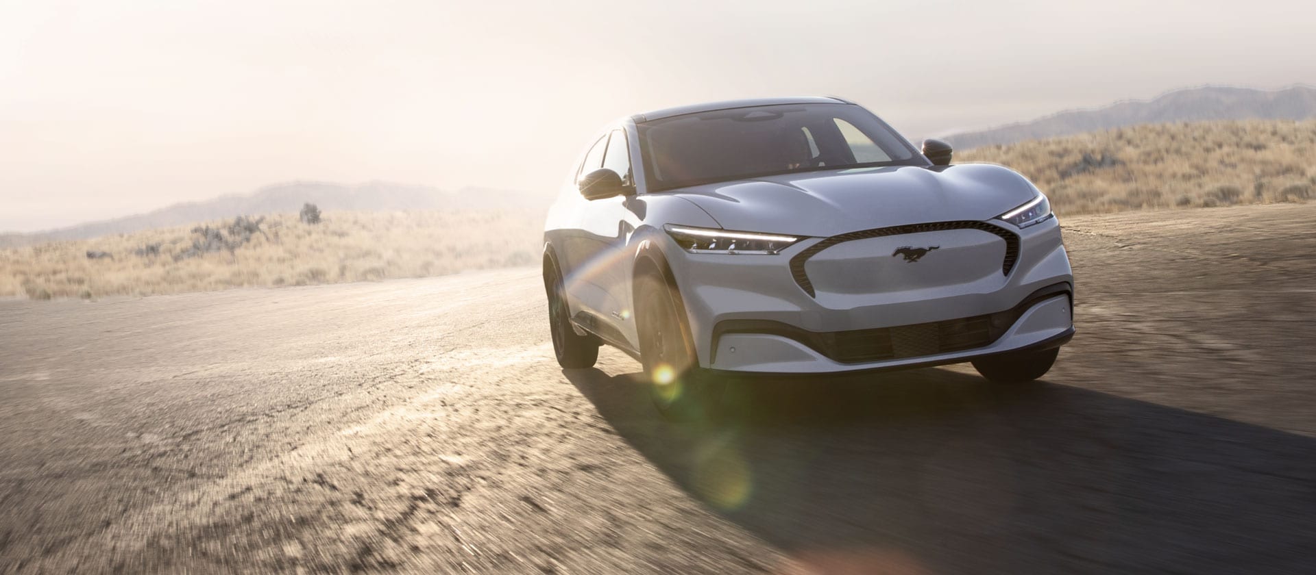 A silver Ford Mustang Mach-E electric vehicle driving on a long open road. 