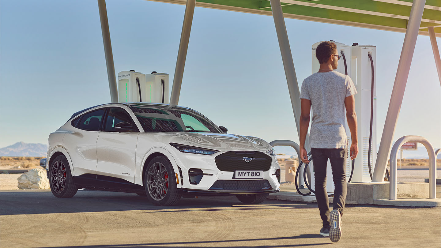 A white Ford Mustang Mach-E parked at an outside public charging station. A man in a white t-shirt walks towards the car.