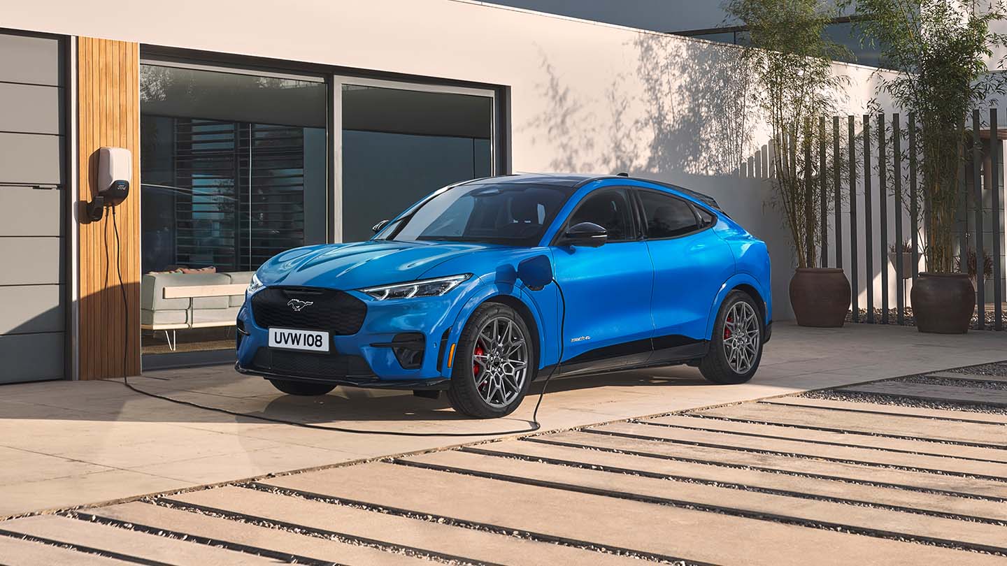 A blue electric Ford Mustang Mach-E plugged into a home charger with a cable while parked on the driveway of a modern house.