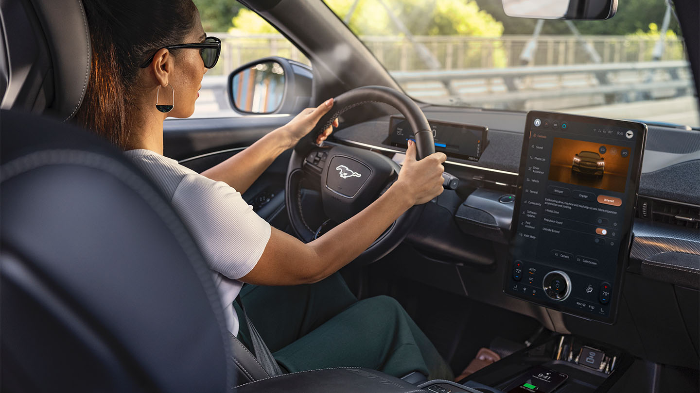 A woman driving the Mustang Mach-E with her hands on the steering wheel. The 15.5" touchscreen shows details about the car.