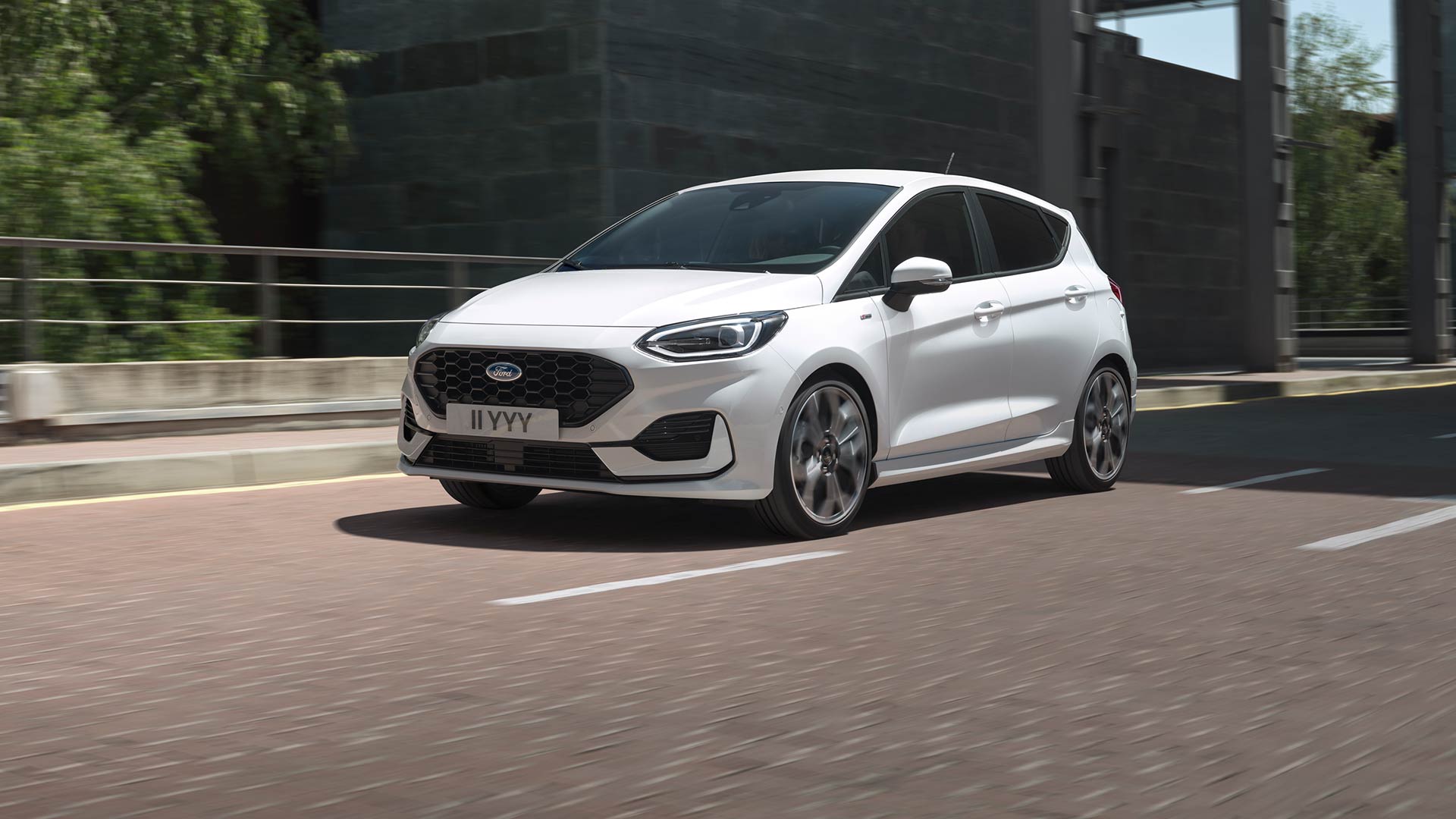 White Ford Fiesta driving on a city road, greenery and buildings in the background.