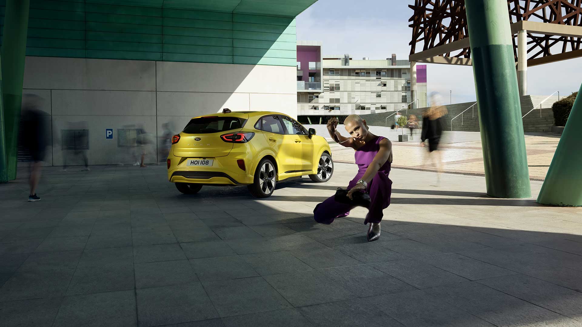 Rear view of a yellow electric Ford Puma Gen-E® parked under a bridge, next to a person dressed in purple.