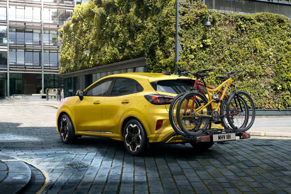 Yellow Ford Puma Gen-E parked in an urban setting with two bicycles mounted on a rear bike carrier.