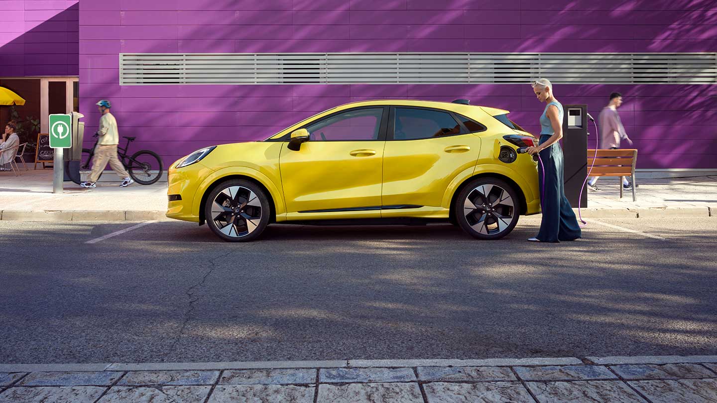 Side view of a yellow electric Ford Puma Gen-E® being plugged into a public charging station while in a parking space.
