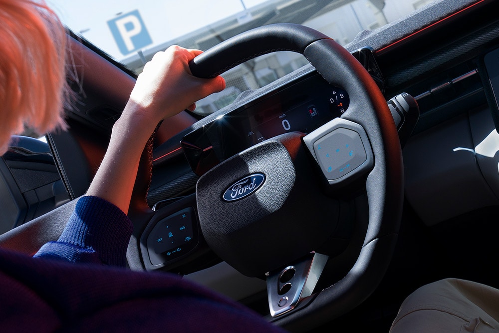 A close-up of a woman holding the steering wheel in a parked Ford Capri, also showing the digital instrument cluster.