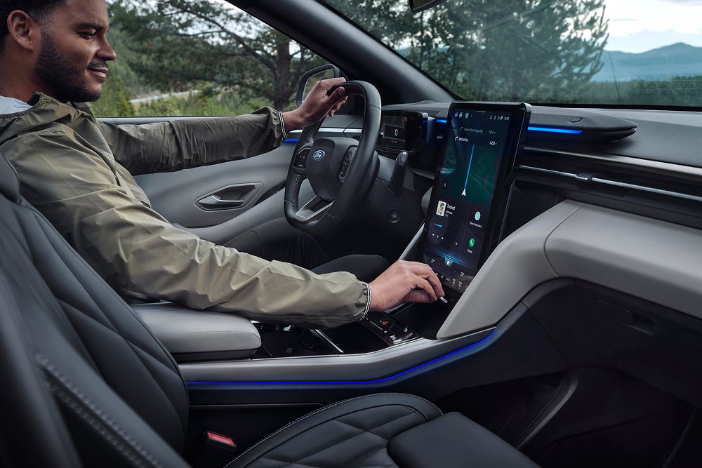 Interior of a Ford Capri with a person driving, showing the steering wheel, large touchscreen, and blue ambient lighting.