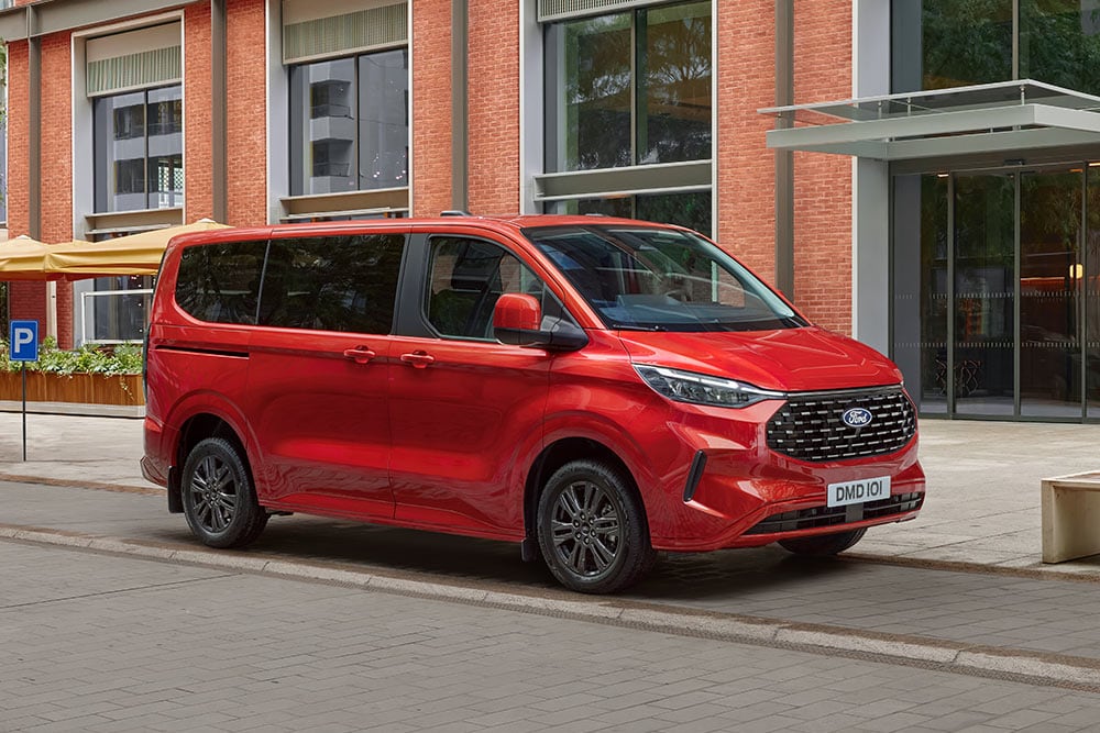 A red Ford Tourneo Custom is parked on a city street in front of a brick building
