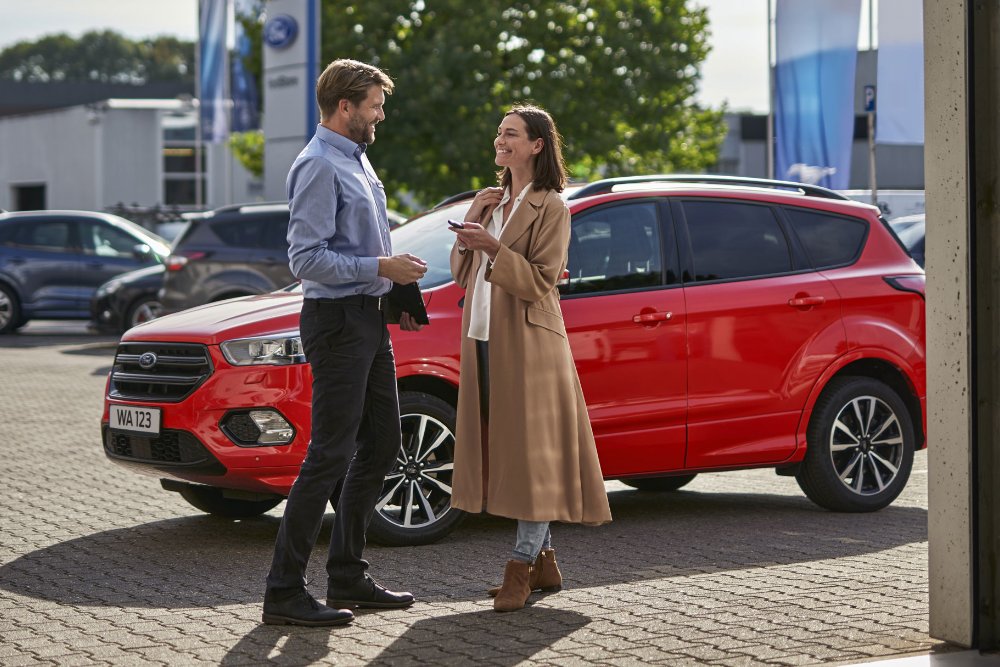 A man and a woman talk next to a red Ford Kuga in a dealership car park.
