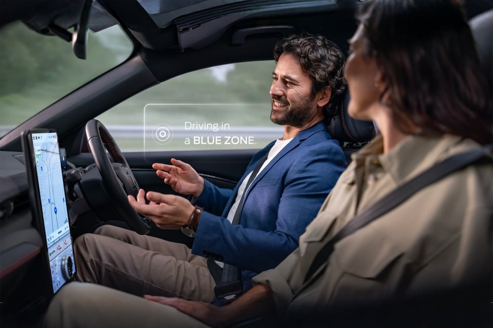 A man and woman in a Mustang Mach-E driving hands-free with Ford BlueCruise. A notification says "Driving in a BLUE ZONE".
