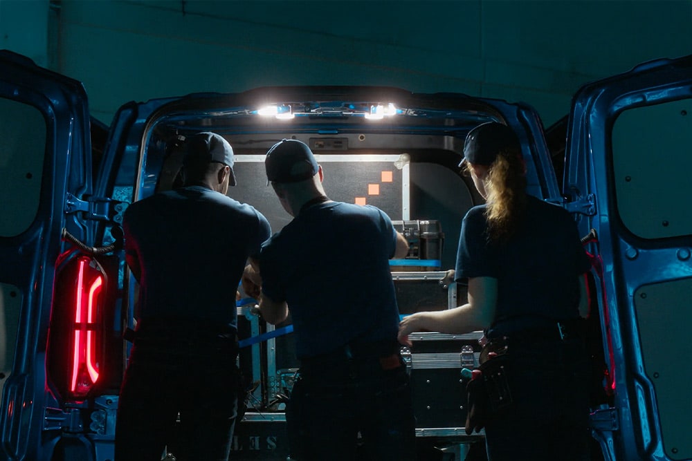Three people working in the back of a Ford Transit Custom van at night, lit by cargo LED lights.