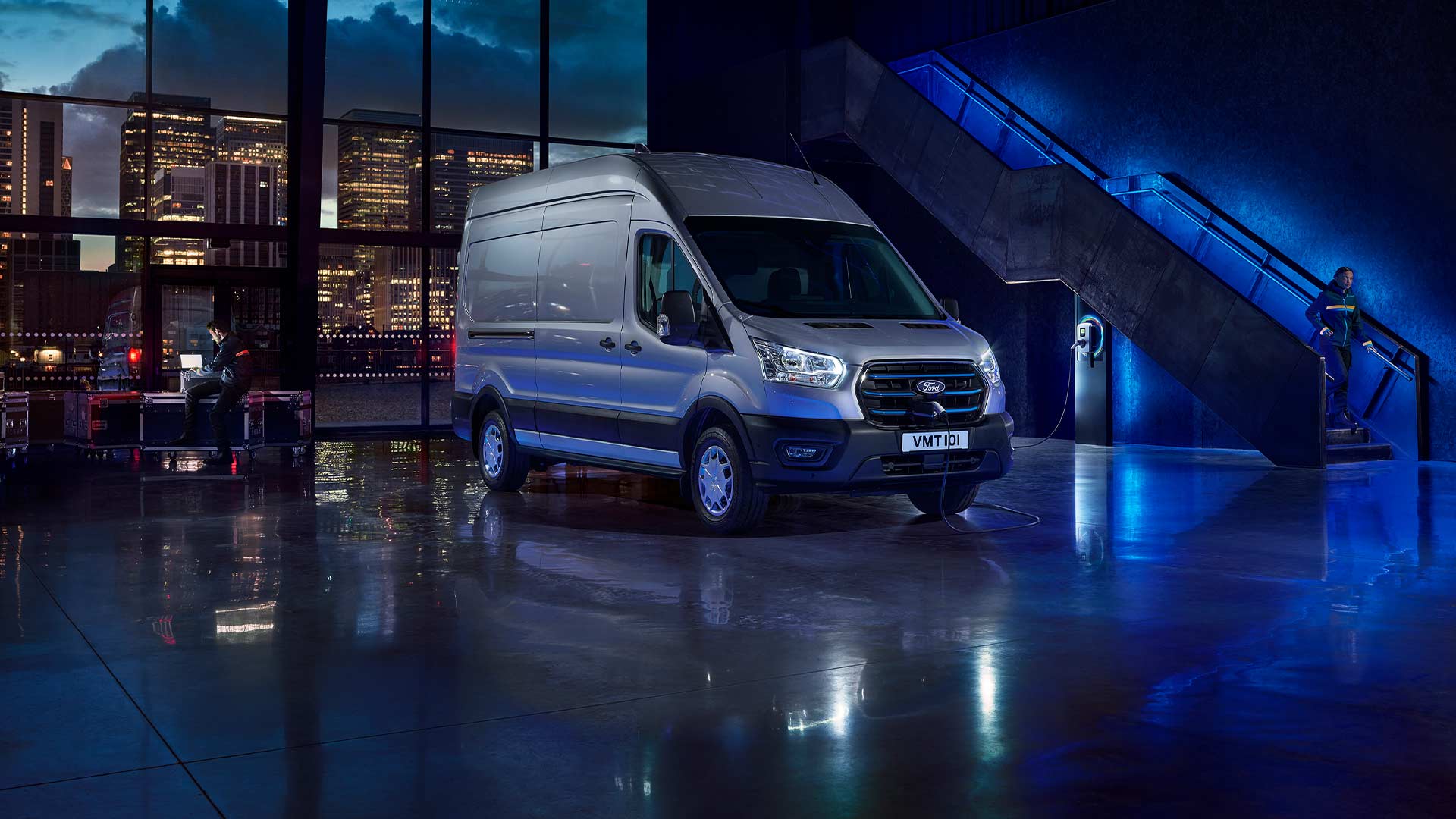 Silver Ford E-Transit electric van parked indoors with a city skyline visible through large windows. 