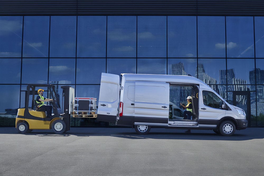  Two people loading cargo into a silver Ford Transit van with a forklift.