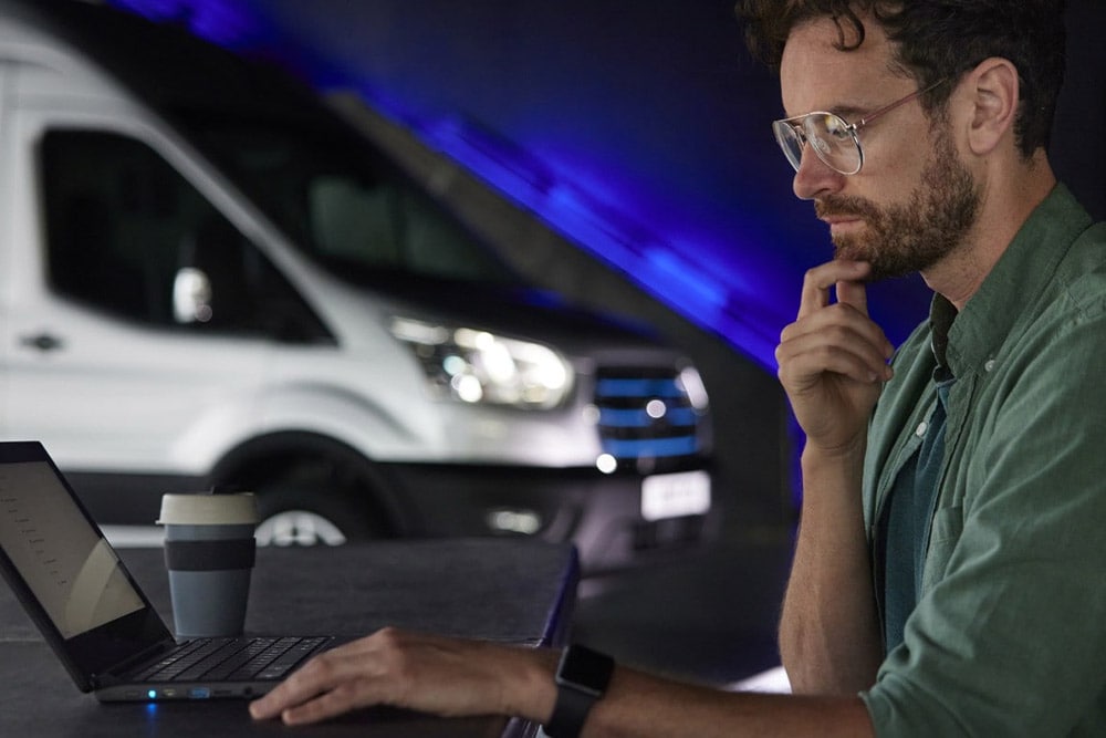 Man working on a laptop with a Ford E-Transit electric van in the background.