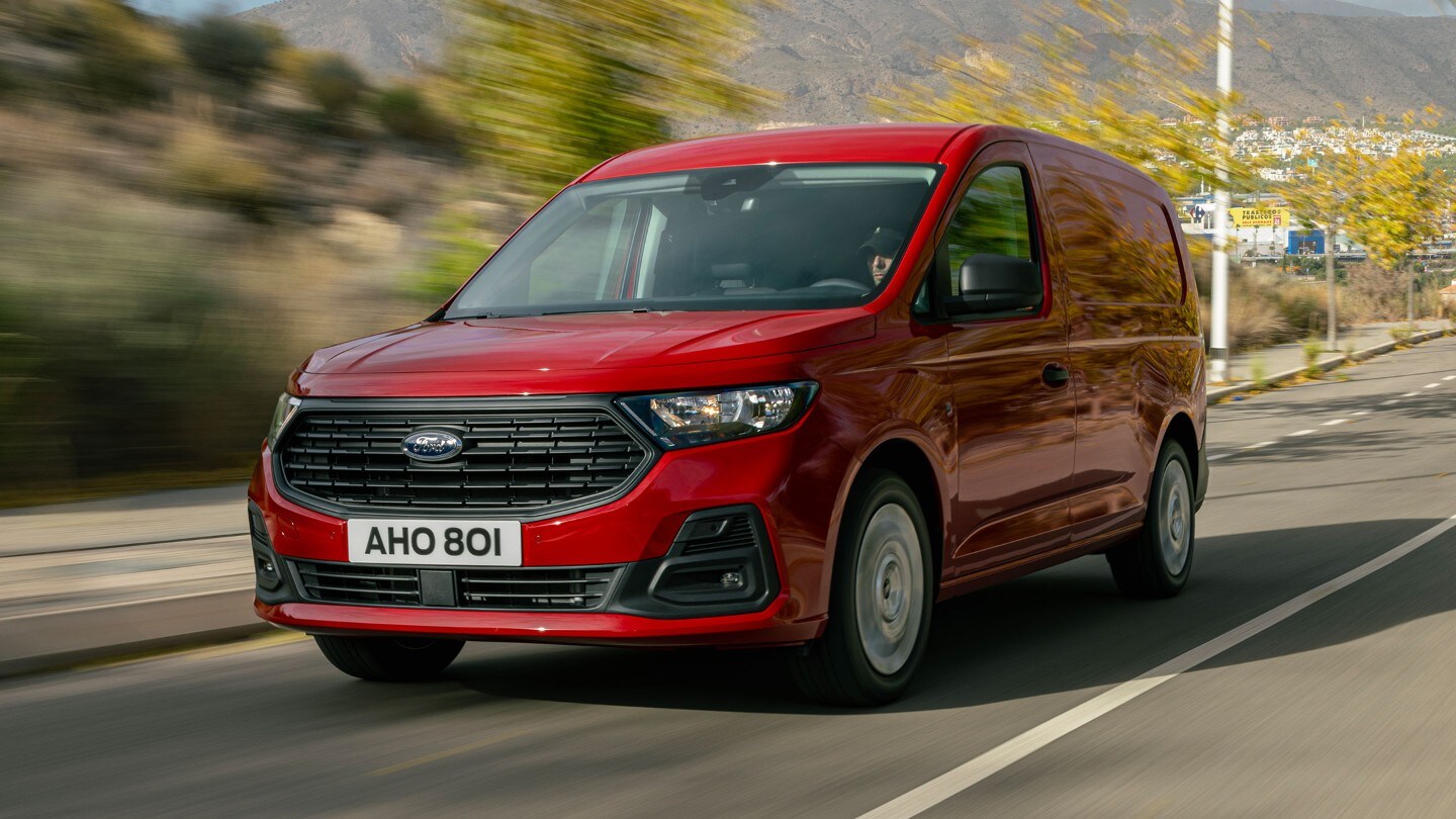 All-New Ford Transit Connect in Maple Red drives along a road showing front grille and headlights. 