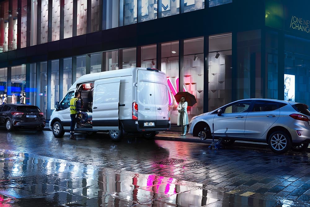White Ford Transit with side door open parked on a raining city street between two cars.
