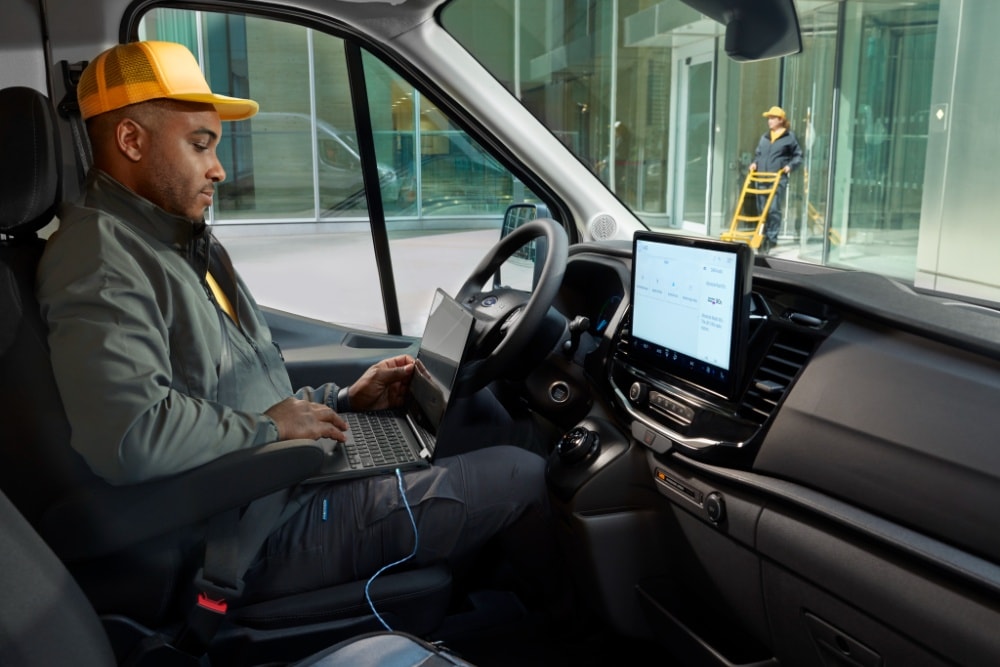 A man sitting in the driver's seat of a Ford Transit van, using a laptop.