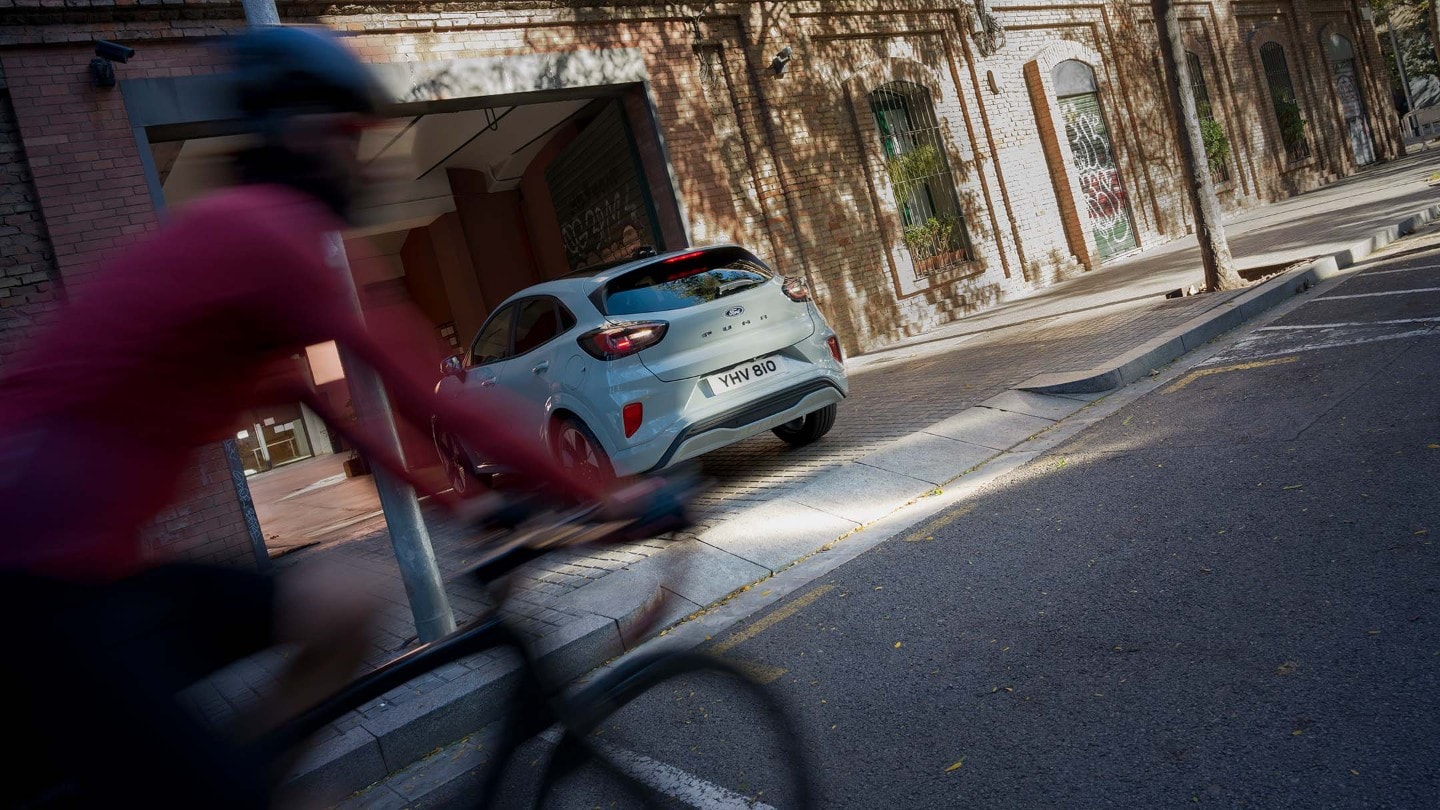 A grey Ford Puma reversing out of a driveway while a cyclist wearing red passes by