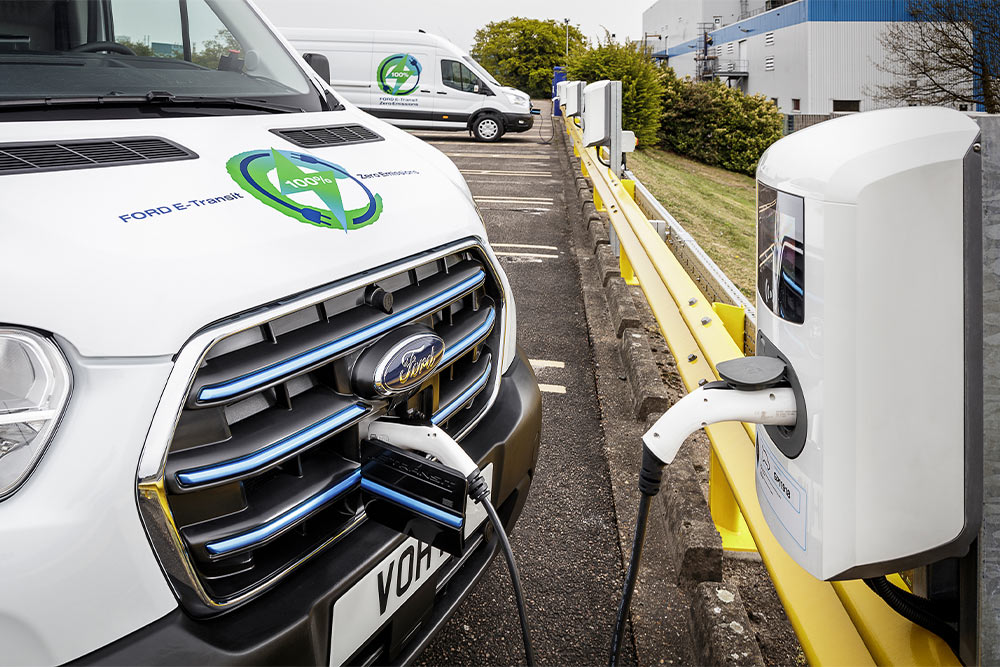 Front view of a white Ford E-Transit van charging at an electric station. A '100% Zero Emissions' logo is on the bonnet.