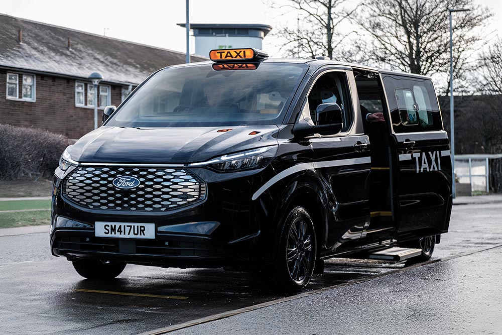 Front 3/4 view of a black Ford Transit Custom taxi with an open side door and a roof-mounted 'TAXI' sign.