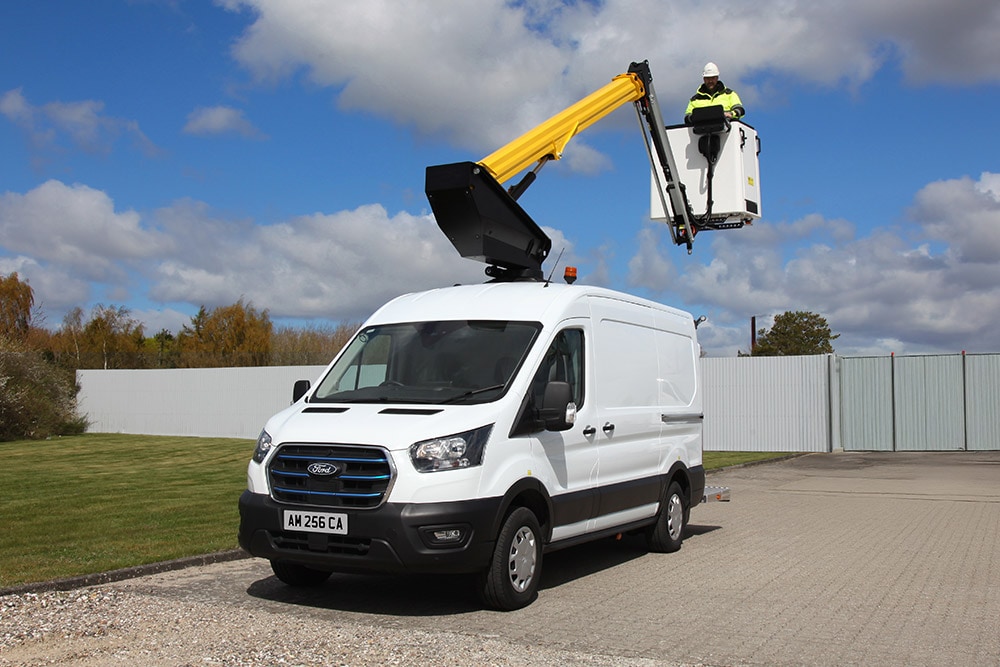 A white Ford Transit van with an extended cherry picker, a technician in the basket, under a blue sky.