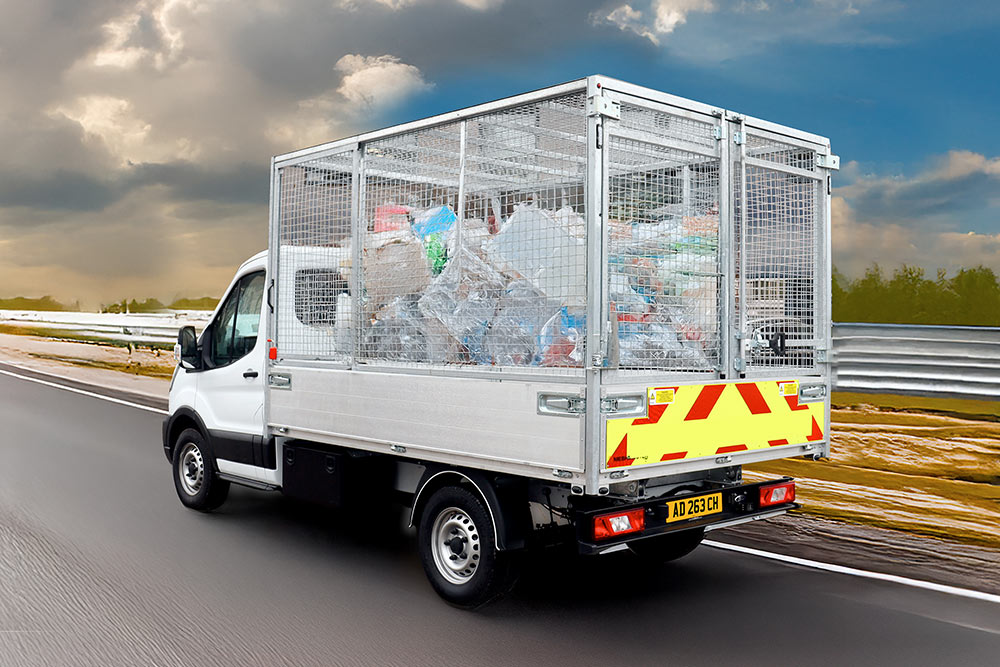 Rear view of a white Ford Transit cage tipper truck, full of rubbish, driving on a road.