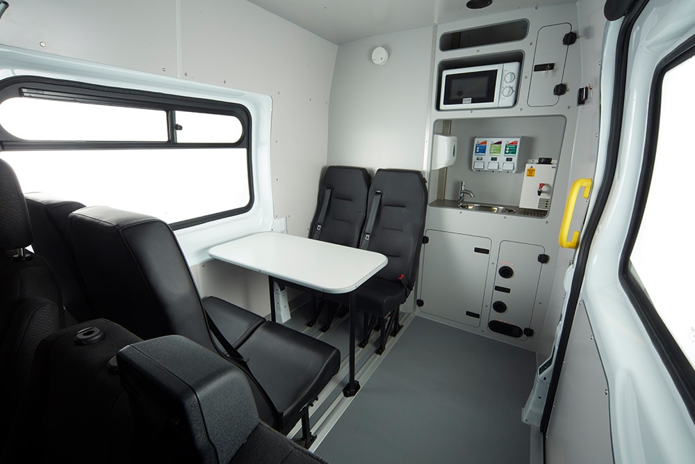 Interior of a Ford van, featuring a table, black seats, and a compact kitchenette with a microwave.
