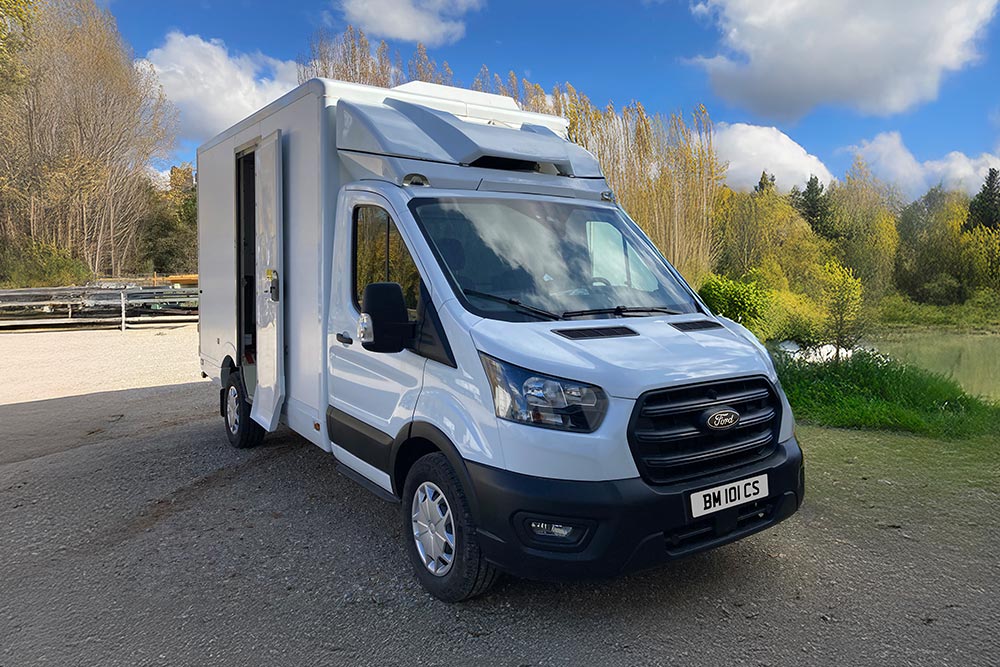 Front 3/4 view of a white Ford Transit box van parked by a lake with trees.