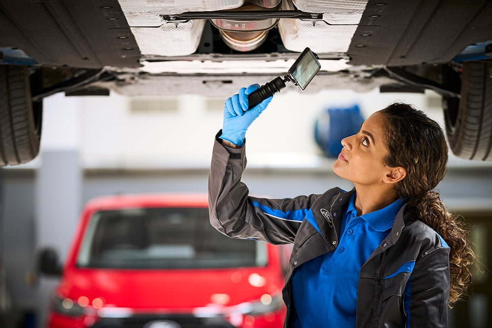A Ford mechanic inspecting the undercarriage of a vehicle, a red Ford van parked in the background.