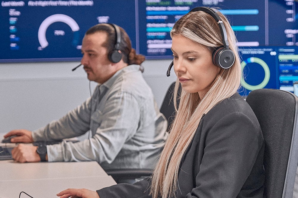 Two individuals with headsets focused on computer screens in a modern control room, with data displays behind them.