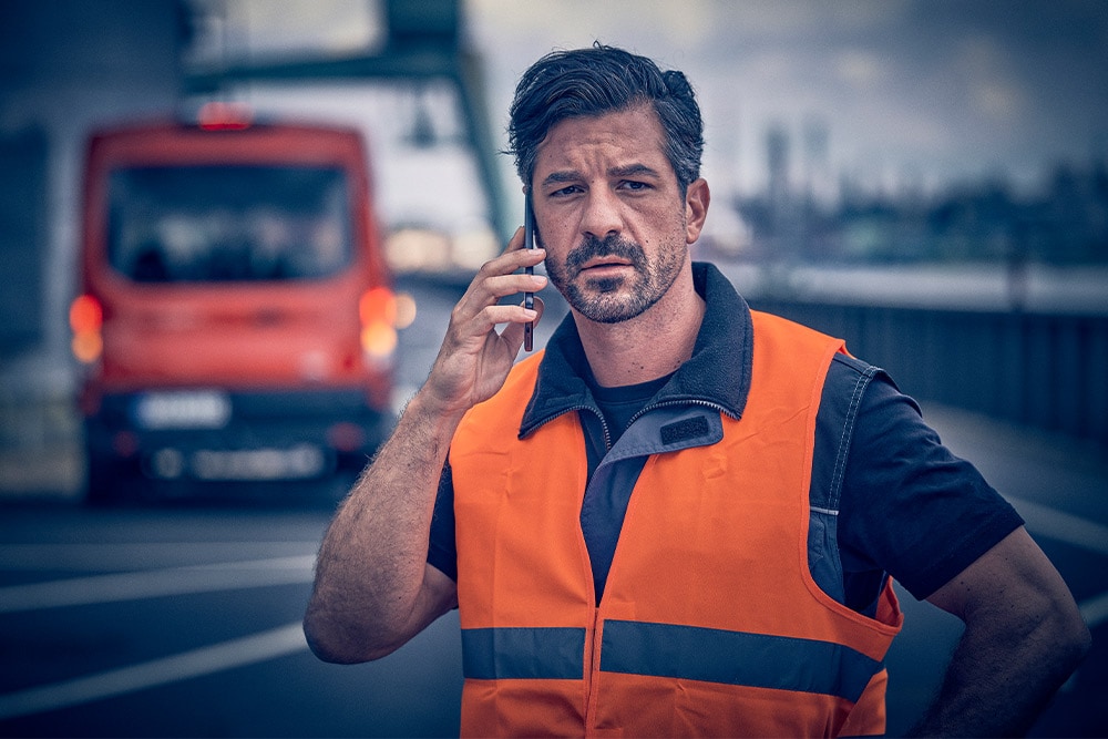 Worker in orange vest calling roadside assistance on a mobile device.