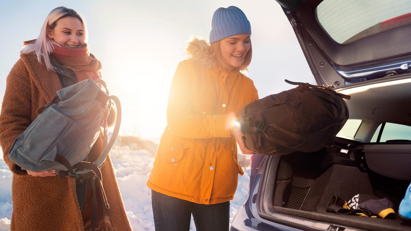 A smiling woman in an orange winter jacket loads a duffel bag into the open trunk of a car in a sunny, snowy setting